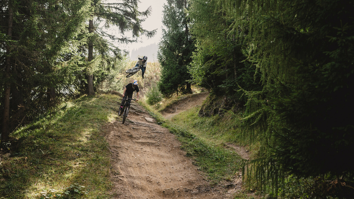 Ein Mountainbiker fliegt in einem Trick hoch in die Luft, während ein anderer Fahrer auf einem Waldweg fährt. Die Szene ist von hohen Bäumen und üppiger Vegetation umgeben.