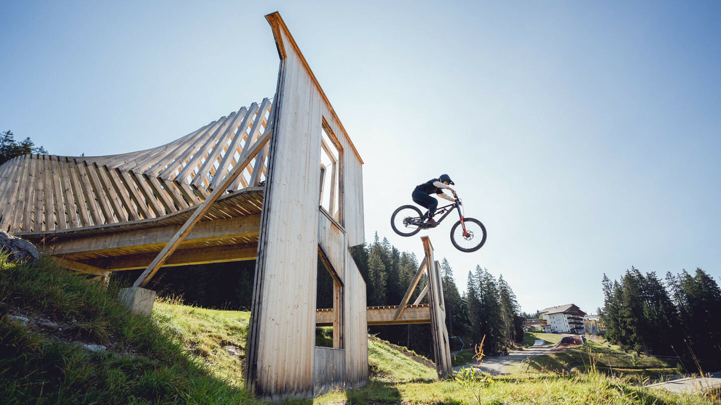 Un cycliste de montagne saute au-dessus d'une rampe en bois dans un parc de vélo. La structure massive en bois se détache sur le ciel dégagé et la nature environnante.