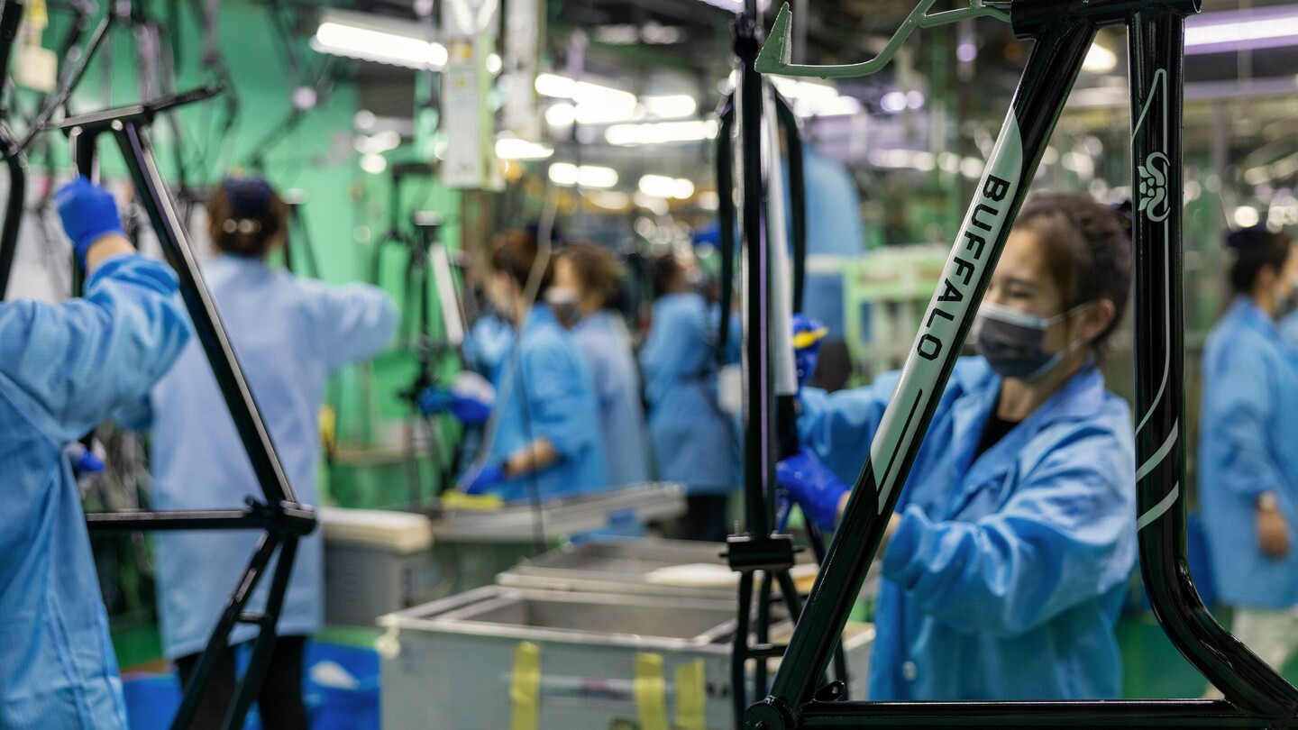 Produktionshalle mit mehreren Personen in blauen Kitteln und Handschuhen, die Fahrradrahmen montieren. Auf einem der Rahmen ist das Logo „BUFFALO“ sichtbar. Die Arbeitsumgebung ist industriell mit vielen Werkzeugen und Beleuchtungen.