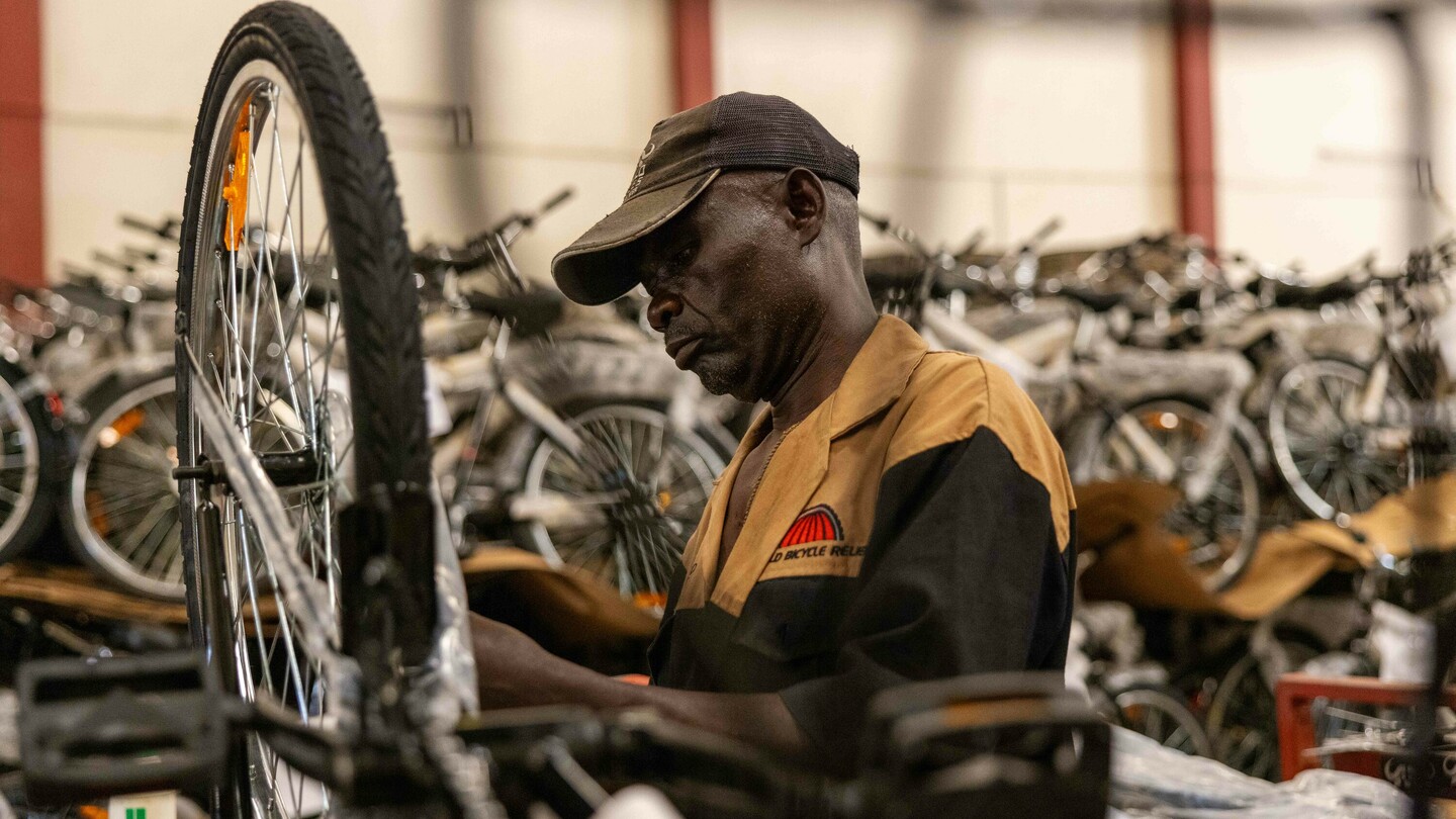 A man in a workshop assembles a bicycle while wearing a cap. Numerous other bicycles are visible in the densely packed background.