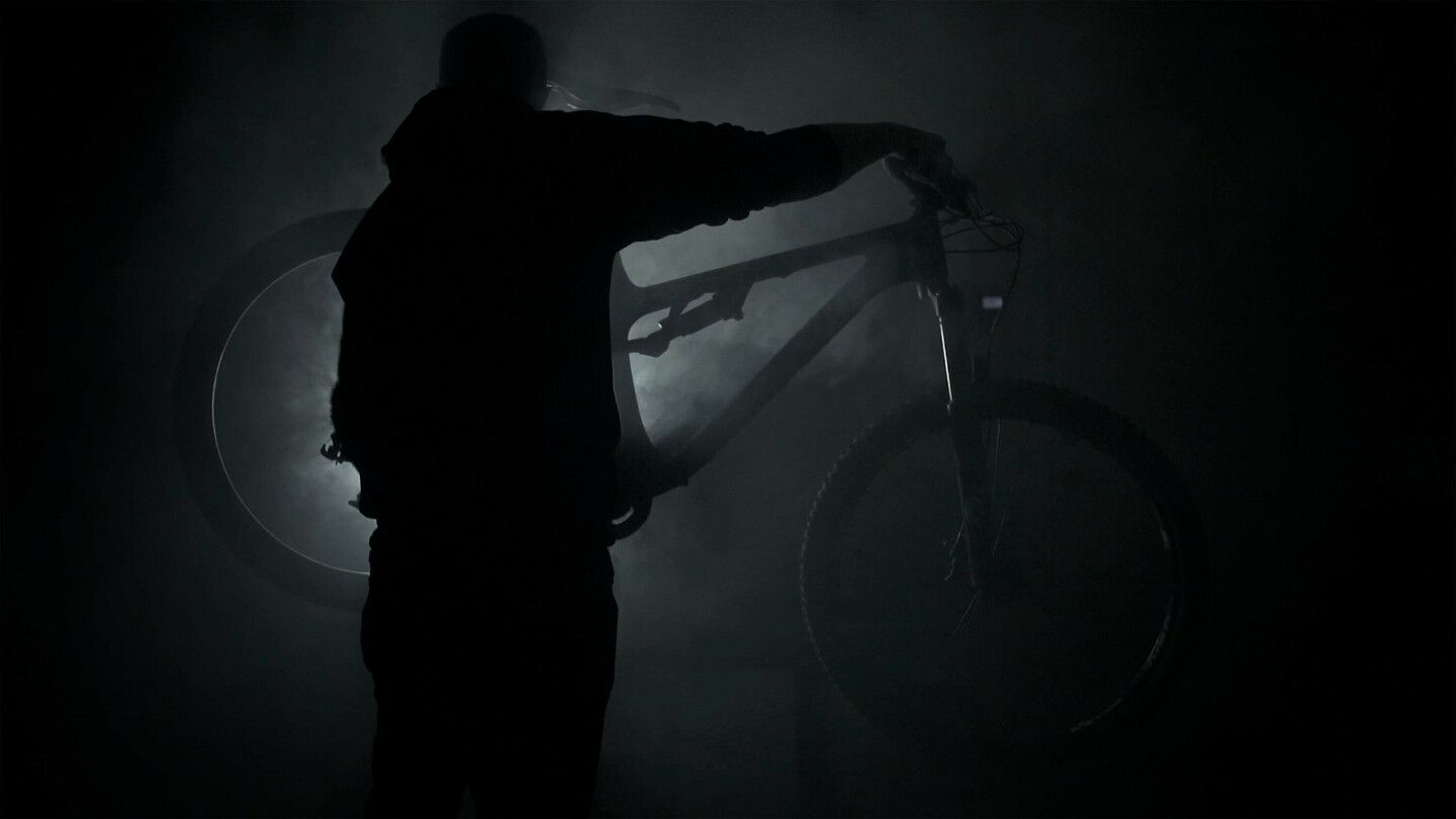 Silhouette of a person holding a mountain bike in a dark, smoky environment with light shining from behind.