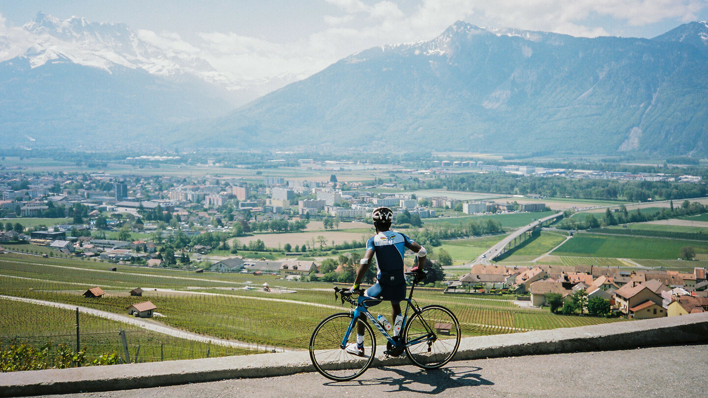 Ein Radfahrer steht neben seinem Fahrrad und blickt auf ein Tal mit einer Stadt, Feldern und Bergen im Hintergrund an einem sonnigen Tag.